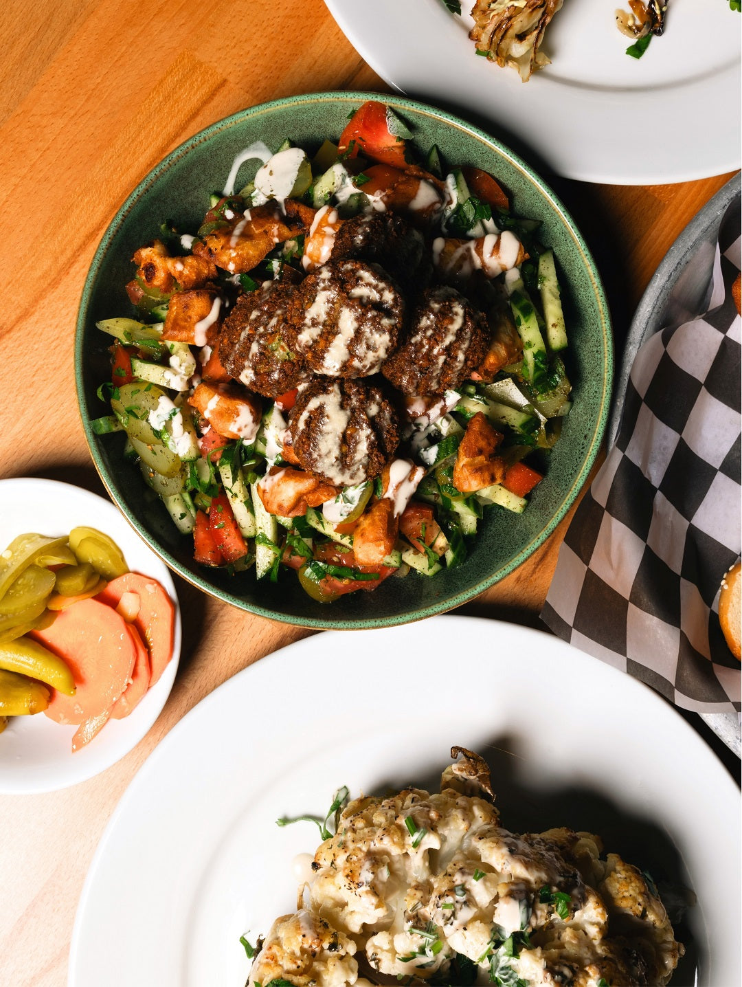 Fresh falafel salad at Mazal Shawarma & Grill topped with tahini, served alongside pickles and roasted cauliflower on a shared table.