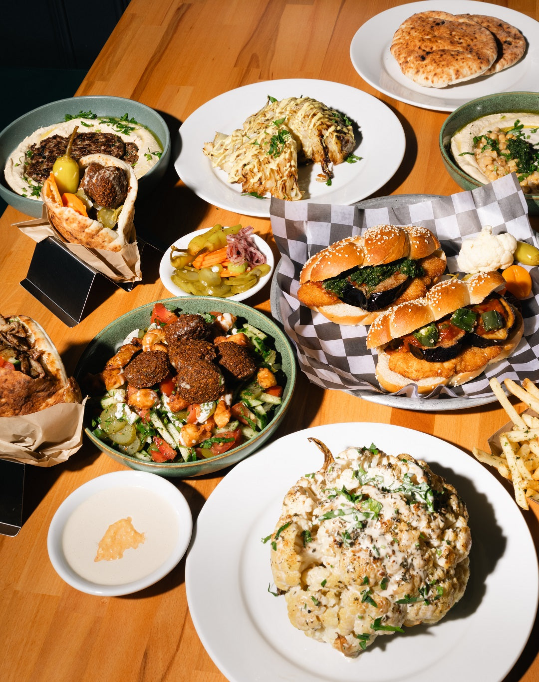 Large table spread at Mazal Shawarma & Grill featuring hummus plates, falafel salad, roasted cauliflower, house baked baguette sandwiches with schnitzel and eggplant, assorted pickles, and fresh pita bread.